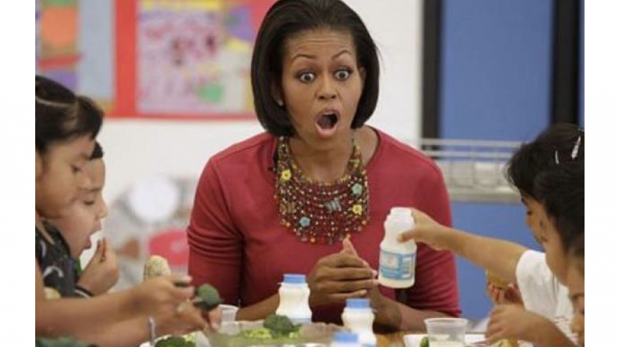 Michelle Obama sitting with school age children eating in a cafeteria. Michelle looking aghast at what the children are eating.